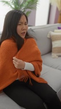 Turuncu bir battaniyeye sarılmış Asyalı bir kadın, gri bir koltukta rahat bir oturma odasında oturuyor, ev ortamında sıcaklık ve rahatlığı resmediyor..