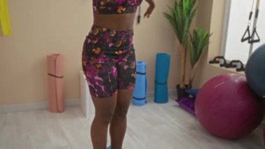 Spor salonunda örülü saç stiliyle esneyen, renkli spor kıyafetleri giyen, spor aletleri ile çevrili bir kadın ve iyi ışıklandırılmış iç dekorasyonda bir bitki..