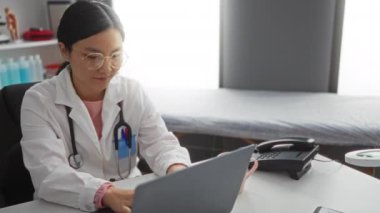 Klinik odasındaki doktor laptopta çalışır. Etrafı tıbbi ekipmanlarla çevrilidir. Odaklanma ve profesyonelliği gösterir..