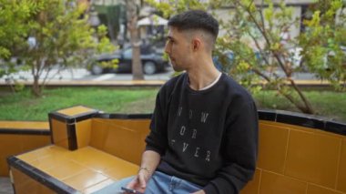 Şehir parkındaki sarı bankta oturan genç adam elinde telefonla düşünceli bir ifadeyle çevresi yeşillikle çevrili.