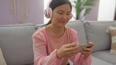 Mobil oyun oynayan bir kadın rahat gri bir kanepeye oturmuş, rahat, parlak bir oturma odasında kulaklık takıyor..