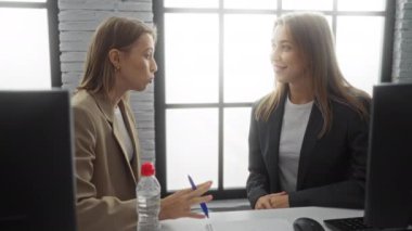 Kadınlar ofis masasında iş konuşuyorlar. Geniş pencerelerden içeri doğal ışık giriyor, takım çalışmalarını ve profesyonel kıyafetleri vurguluyorlar..