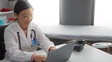 Klinik masasında çalışan kadın doktor, dizüstü bilgisayar kullanıyor, heyecan gösteriyor, arka planda tıbbi ekipmanlar var, modern bir sağlık hizmeti sunuyor..