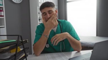 Yeşil önlüklü doktor hastane ofisindeki bir masaya dizüstü bilgisayarı ve tıbbi ekipmanlarla çevrili evrak işleriyle ilgileniyor..