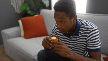 Genç adam oturma odasında mum kokusu alıyor, kanepede çizgili gömleğiyle rahatça oturuyor, evde dinleniyor.