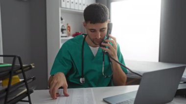 Yeşil önlüklü genç bir adam hastane odasında belgeleri incelerken telefonla konuşuyor, profesyonel bir sağlık hizmeti sunuyor..