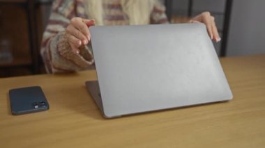Konforlu oturma odasında dizüstü bilgisayar kullanan bir kadın, iç mekandaki ekrana odaklanmış, gündelik kazaklı sarışın bir kadını gösteriyor..