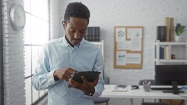 Ofisteki genç adam tablet kullanırken gülümsüyor, modern dekorasyon ve doğal ışık ile çevrili, profesyonel ve ilgi çekici bir çalışma ortamı yaratıyor..