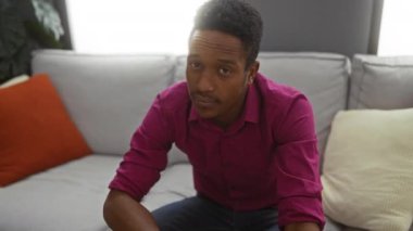 Oturma odasında kafasına dokunan pembe gömlekli genç bir adam, gri bir kanepede oturan, iç mekan renkli yastıklı, düşünceli, yetişkin bir erkeği canlandırıyor..