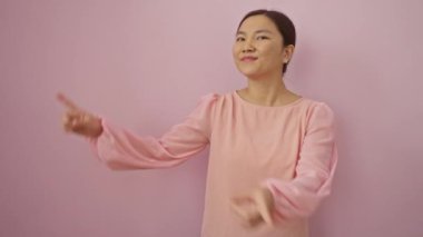 Gülümseyen genç Çinli kadın iki eliyle yalnız pembe arka planı işaret ederek kameraya güvenle bakıyor.