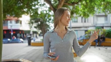 İki gülümseyen kadın, arka planda ağaçlar ve binalar olan bir şehir parkında arkadaşlık, mutluluk ve modern yaşam tarzını sergileyerek selfie çekiyorlar..