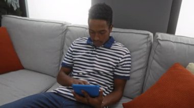 Tablet kullanan bir adam, rahat bir şekilde evdeki bir kanepede oturuyor. Günlük çizgili gömlek giyiyor ve gülümsüyor. Modern bir oturma odasında rahatlama ve rahatlama öneriyor..