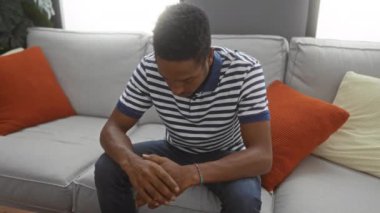 Genç adam rahat bir oturma odasında, renkli yastıklarla çevrili çizgili gömleği ve düşünceli ifadesiyle rahat bir iç mekan atmosferi yaratıyor..