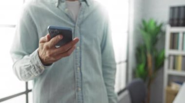 Ofisteki genç adam, arka planda büyük pencereler ve yeşilliklerle akıllı telefondan mutlu bir şekilde konuşuyor. Profesyonel bir çalışma ortamı ve olumlu bir iletişim öneriyor..