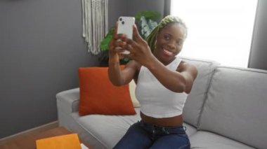 Örgülü kadın gülümsüyor ve rahat oturma odasında akıllı telefon kullanıyor. Arka planda turuncu aksanlı yeşil bitki var. Sıcak ve davetkar bir atmosfer yaratıyor..