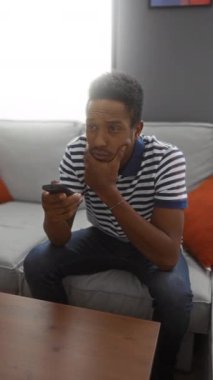 Modern oturma odasındaki kanepede uzanan, çizgili gömlek giyen, uzaktan kumandalı, parlak doğal ışık ve gündelik ev düzeniyle düşünceli görünen bir adam..