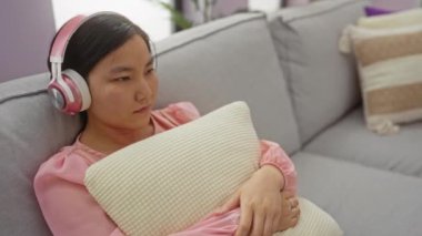 Kulaklıklı bir kadın yastığı tutuyor rahat bir evde rahat bir koltukta dinleniyor pembe bir gömlek giyiyor ve müzik dinliyor, huzurlu ve rahatlatıcı bir oturma odası atmosferi yaratıyor..