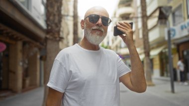 Gri saçlı ve sakallı olgun bir adam şehir sokağında dikilirken telefonundaki sesli mesajı dinliyor.