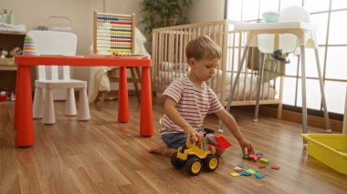 Sevimli, sarı saçlı, yerde oyuncak kamyonuyla oynayan sevimli bir çocuk. Etrafı renkli oyuncaklar ve mobilyalarla çevrili, kapalı bir anaokulu ortamında.