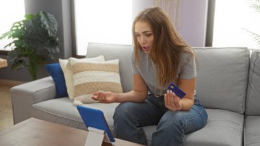 Bir video konferansına katılan bir kadın, elinde modern mobilyalarla rahat bir oturma odasında kredi kartı tutarak, gündüz vakti evde bir ev ortamını sergiliyor..