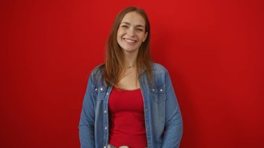 Canlı kırmızı bir arkaplan karşısında gülümseyen kadın, rahat ve kendinden emin bir tavırla kot giymiş, izole bir ortamda genç ve çekici bir görünüm sergiliyor..