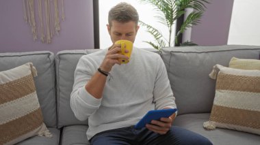 Genç adam oturma odasında bir kanepede oturuyor, elinde bir tablet tutuyor ve sarı bardaktan içiyor, evinde rahat ve modern bir hayat sürüyor..