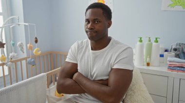 Bebek beşiği ve oyuncaklarıyla rahat bir yatak odasında kollarını kavuşturan yakışıklı bir adam, ev ortamında sükunet ve memnuniyeti yansıtıyor..