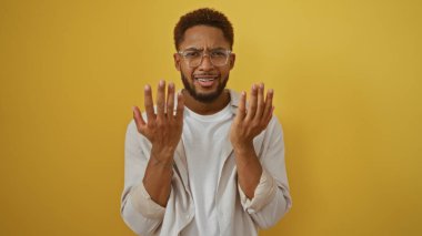 Sarı arka planda duran genç adam elleri havada, gözlük takıyor ve sıradan bir kıyafet giyiyor..