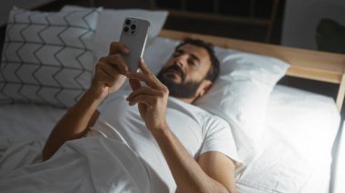 Yakışıklı İspanyol adam, gece vakti rahat bir yatak odasında yatarken akıllı telefon kullanıyor.