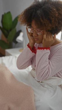Kıvırcık saçlı bir kadın yatak odasındaki bir yatağa oturur, baş parmaklarını birbirine bastırır, baş ağrısına işaret eder ve evde huzurlu bir şekilde pijama giyer..