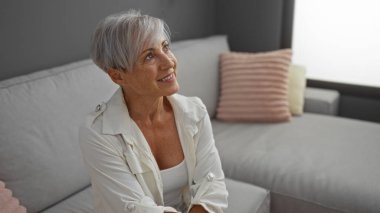 Gri saçlı güzel kadın, kısa saçlı, modern bir oturma odasında gülümseyen, yumuşak ışıklandırmalı, rahat dekorlu, ev ortamında neşe ve zarafet saçan bir kadın..