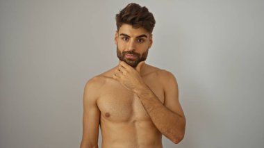 Yakışıklı, sakallı, genç bir adam. Düşünceli bir şekilde çenesine dokunuyor. Beyaz bir arka plan duvarından izole edilmiş..