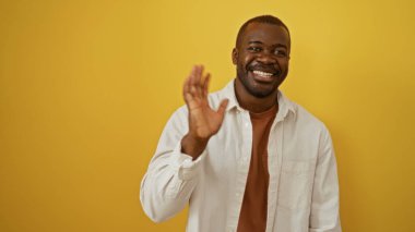 Yakışıklı Afro-Amerikalı genç adam, parlak sarı bir arka plana el sallarken sıcak bir şekilde gülümsüyor.
