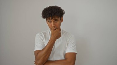 Beyaz tişörtlü yakışıklı İspanyol bir adam beyaz bir arka planda, düşünceli bir ifadeyle tek başına duruyor.