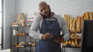 Genç adam fırında tablet kullanırken telefonla konuşuyor ve arka planda ekmek ve hamur işleri var.