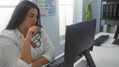 Bir ofis odasında çalışan, elinde gözlük olan ve bilgisayar ekranına bakan İspanyol bir kadın.