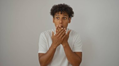 Beyaz tişörtlü kıvırcık saçlı genç adam elleri beyaz arka planda ağzını kapatırken şaşırmış görünüyor.