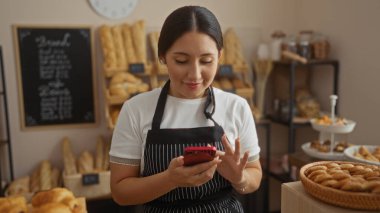Kadın fırında cep telefonu kullanıyor, arka planda ekmek ve hamur işleri var, siyah önlük giyiyor, ifadesini odaklıyor, esmer saçlı, İspanyol görünümlü, genç, kadın, ev ortamında.