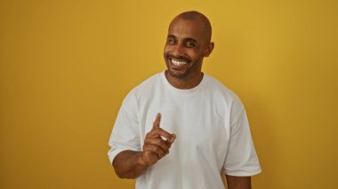 Yakışıklı, Afro-Amerikalı, izole edilmiş sarı arka planda gülümseyen, beyaz gömlekli, tek parmakla işaret eden bir adam.