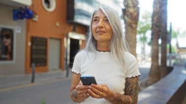 Gri saçlı, elinde akıllı telefon olan yaşlı bir kadın arka planda palmiye ağaçları ve modern binalar olan bir şehir sokağında dikiliyor..