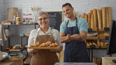 Kadın ve erkek, sıcak kapalı bir fırında taze kruvasanlarla gururla poz vererek ekmek ve hamur işi dolu bir dükkanda fırıncı ve ortak olarak çalışmalarını sergiliyorlar..