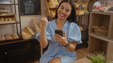 Fırında kutlama yapan bir kadın akıllı telefonu kontrol ediyor, iyi haber ve başarıdan mutlu, ekmek ve pastalarla çevrili..