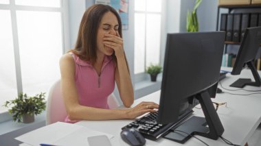 Ofis ortamında bilgisayarın başında esneyen genç bir kadın, yorgun ya da uykulu görünüyor, iş yerinde bitkinlik ya da yorgunluk öneriyor, görünür bitkilerle iç aydınlatma altında