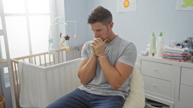 Genç adam düşünceli bir şekilde bir beşikteki parlak bir çocuk odasında oturuyor, ebeveynliği yansıtıyor, bebek gereksinimleriyle çevrili, kapalı bir odada..
