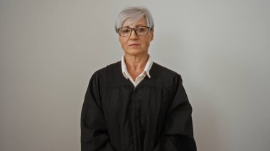 Olgun, gri saçlı, siyah cüppeli kadın yargıç ciddi bir ifadeyle beyaz bir arka plan duvarına yaslanmış duruyor..