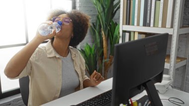 Ofis odasında su içen kadın masasında oturan kıvırcık saçlı iş ortamını ve su ihtiyacını gösteren bir kadın..
