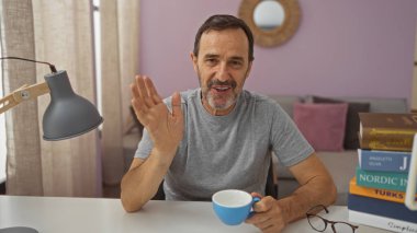 Latin asıllı orta yaşlı bir adam, elinde bir bardak tutarken kameraya el sallıyor. Elinde bir lamba ve masada kitaplar var..