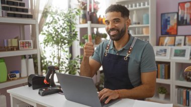 Önlük giymiş gülümseyen adam arka planda bitki ve raflar olan bir ev dekoratöründe baş parmak işareti yapıyor.