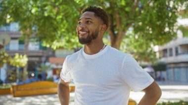 Arka planda yeşillik ve binalar olan bir şehir parkında güvenle gülümseyen genç adam.