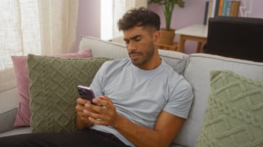 İspanyol bir adam rahat bir oturma odasındaki kanepeye uzanmış, akıllı telefonuna bakıyor, düşünceli bir ifadeyle, yastıklarla ve ev bitkileriyle çevrili..